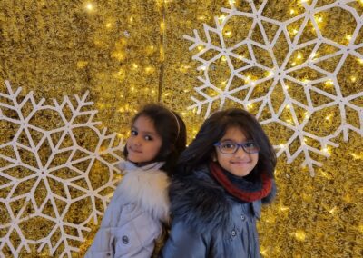 Riya and her sister Niki wearing warm coats. They smile at the camera in front of a winter display with white snowflakes