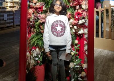 Riya is standing in a converted red phone box and surrounded by red, white and pink flowers